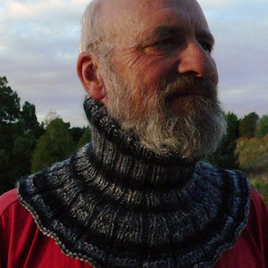 Op de afbeelding: Een man draagt een zwarte en grijze gebreide cowl neck sjaal. De sjaal is dik en heeft een gegolfde rand.