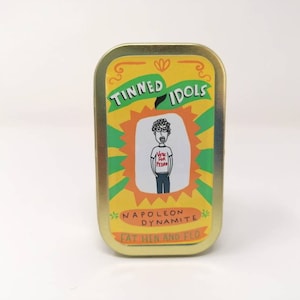 Könnte beinhalten: Eine gelbe, grüne und orangefarbene Dose mit den Worten "Tinned Idols" und "Napoleon Dynamite" darauf gedruckt. Die Dose zeigt eine Comic-Illustration einer Person, die ein T-Shirt mit der Aufschrift "Vote for Pedro" trägt.