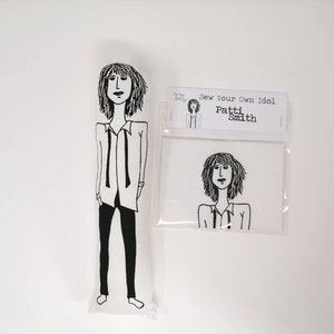 May include: Black and white line drawing of Patti Smith on a white background. The drawing is on a white fabric doll with the text "Sew Your Own Idol Patti Smith" on a white card.