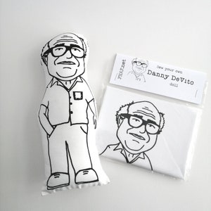 May include: A black and white line drawing of Danny DeVito on a white fabric doll. The doll is ready to be colored in. The packaging includes a smaller version of the drawing on a white background with the text "Sew your own Danny DeVito doll" and the website "THAF.net".