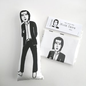 May include: Black and white illustration of a person on a white pillow. The illustration is of a person with black hair and a black suit. The pillow has the text "Sew your own Nick Cave doll" printed on it.