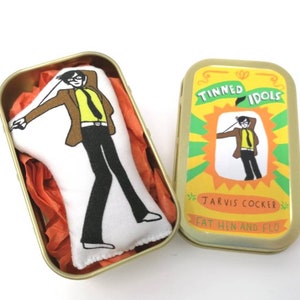May include: A small tin box with a yellow background and a cartoon image of a man with a microphone. The text on the tin reads "Tinned Idols Jarvis Cocker Fat Hen and Flo".
