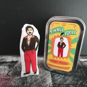 Può includere: Una scatola di latta con sfondo giallo e accenti verdi. Il testo "Tinned Idols" è stampato in lettere nere. Un'immagine di un uomo con baffi e barba in stile cartone animato è all'interno della scatola. Il testo "Pedro Pascal" è stampato sotto l'immagine.