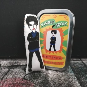 May include: A colorful tin with the text "Tinned Idols" and "Robert Smith" on it. The tin features a cartoon image of Robert Smith, the lead singer of The Cure. The tin is next to a plush toy of Robert Smith.