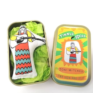 May include: A yellow, green, and orange tin with the text "Tinned Idols Joni Mitchell Fat Hin and Flo" and a cartoon image of a woman playing a guitar. The tin contains a small, white fabric doll of a woman playing a guitar.
