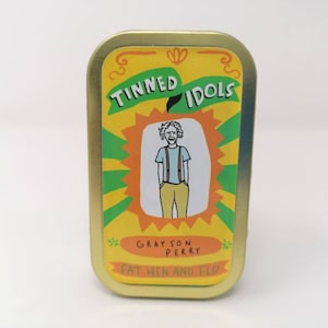 Op de afbeelding: Een gele en groene tinnen doos met de tekst "Tinned Idols Grayson Perry Fat Hen and Flo" en een cartoonillustratie van een persoon die een blauw shirt, bretels en een gele broek draagt.
