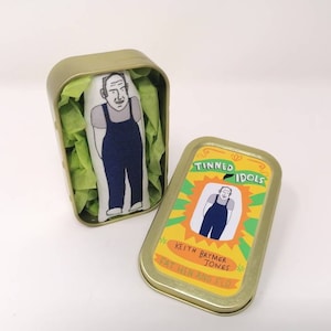 May include: A small tin box with a yellow and green label that says "Tinned Idols" and features a cartoon illustration of a person wearing overalls. The text on the label also reads "Keith Brymer Jones Eat, Drink and Flourish".