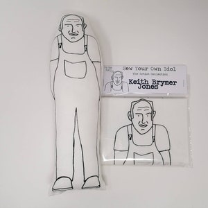 Op de afbeelding: Een zwart-witte lijntekening van een persoon die een overall draagt. De tekening is op een wit stoffen kussen. Het kussen is verpakt in een plastic zak met een label waarop staat "Sew Your Own Idol The Artist Collection Keith Brymer Jones".