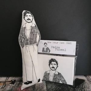 May include: A white fabric doll of a person with black hair and a black and white checkered shirt. The doll is holding a small white card with the text "Sew Your Own Idol Pedro Pascal".