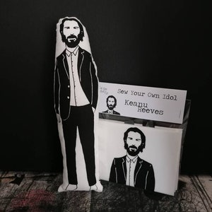 May include: A black and white illustration of Keanu Reeves on a white pillow. The pillow is packaged in a clear plastic bag with a white label that reads "Sew Your Own Idol Keanu Reeves".