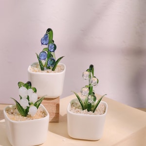Peut inclure: Trois petits pots carrés en céramique blanche contiennent chacun une sculpture de fleur en verre. Les fleurs sont dans des tons de bleu et de blanc, avec des tiges et des feuilles vertes. Les pots sont remplis de petits cailloux clairs.
