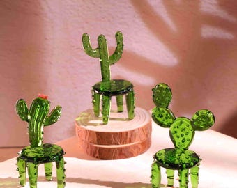 May include: Three decorative glass cactus sculptures in varying designs. The cacti are a vibrant green with small details, including a red flower on one. Each cactus is designed as a chair, with a round seat and four legs.