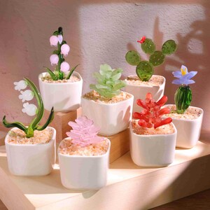 May include: A collection of glass plant sculptures in small white square pots. The plants include a pink flower, a green succulent, a red succulent, a cactus, and a blue flower. The plants sit on a wooden shelf.