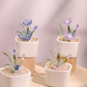 Könnte beinhalten: Vier kleine quadratische weiße Keramiktöpfe, die jeweils ein Glasblumenarrangement enthalten. Die Blumen sind in Blau-, Lila- und Klartönen gehalten, mit grünen Glasstielen und Blättern. Die Töpfe sind mit kleinen, hellfarbigen Kieselsteinen gefüllt.