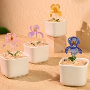 May include: A collection of small, white square ceramic pots, each containing a decorative, colorful flower. The flowers are made of translucent material in shades of pink, purple, yellow, and blue. The pots are filled with small pebbles.
