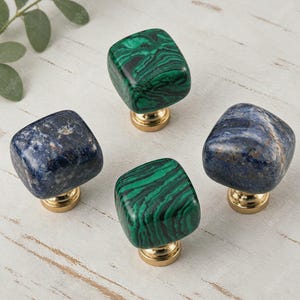 Pode incluir: Quatro puxadores quadrados para armários com topos de pedra polida em padrões de malaquita verde e sodalita azul profundo. Cada puxador é montado numa base metálica circular de tom dourado, exibindo veios minerais naturais.