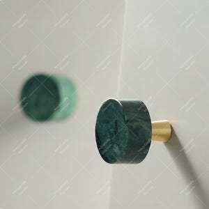 May include: A green marble cabinet knob with a gold-colored base. The knob is cylindrical with a flat, circular face. The marble has dark green veining. The knob is mounted on a white surface.