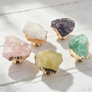 May include: Five cabinet knobs featuring raw, irregular gemstone tops in shades of pale pink, white, deep purple, pale yellow, and light green. Each stone is accented with a metallic gold-toned base and a textured gold-toned edge trim.