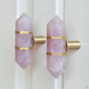 May include: Two rose quartz crystal cabinet knobs with gold wire and brass fittings. The light pink knobs are mounted on a white surface. Each knob is approximately 10 cm long.
