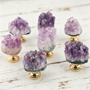 May include: Six cabinet knobs featuring raw amethyst crystal clusters in varying shades of purple and white, each mounted on a polished gold-tone metal base. The natural crystal formations display jagged, translucent points and dark grey rocky bases.