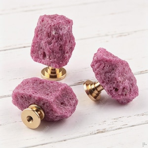 May include: Three cabinet knobs featuring raw, textured magenta-colored stone tops attached to circular, polished gold-tone metal bases. Each knob has a distinct, irregular crystalline shape with a porous surface, mounted on a short metal stem.
