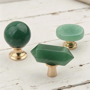 May include: Three cabinet knobs featuring polished green stone tops in spherical, hexagonal, and flat circular shapes. Each piece is mounted on a brushed gold-toned metal base, providing a distinct contrast between the smooth stone and the metallic hardware.