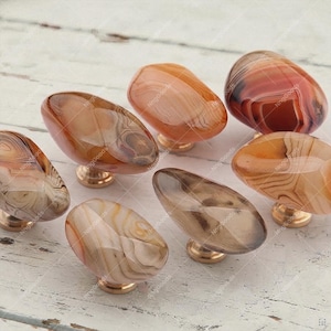 May include: A collection of six cabinet knobs featuring polished, smooth, oval-shaped stones with natural banded patterns in shades of brown, tan, cream, and orange. Each stone is securely mounted on a small, circular, metallic gold-toned base.