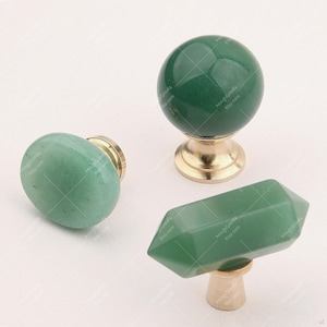 May include: Three green gemstone cabinet knobs in different shapes: an oval, a sphere, and a faceted crystal. Each knob has a brass base. The knobs are displayed on a white surface.