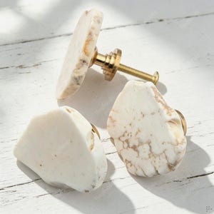 Peut inclure: Trois boutons de placard dotés de faces en pierre blanche de forme organique irrégulière avec des veines fauves. Chaque bouton est fixé à une base en métal doré avec une tige filetée dépassant à l'arrière pour le montage sur meubles.