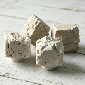 Puede incluir: Cuatro pomos cuadrados para armarios hechos de piedra porosa de color beige claro. Cada pomo presenta una textura rugosa y natural con hendiduras visibles y una forma cúbica, mostrando un acabado crudo y sin pulir en todos sus lados con una pequeña base circular.