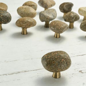 Puede incluir: Una colección de pomos para armarios con piedras de río naturales, lisas y redondeadas, en varios tonos de gris, marrón y beige. Cada piedra está montada de forma segura sobre una base cónica de color latón pulido, creando un herraje funcional.