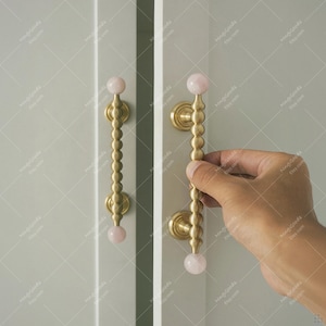 Könnte beinhalten: Schranktürgriffe in Messingoptik, jeweils mit einer Reihe abgerundeter Details und einer rosa Kugel am Ende. Das Bild zeigt eine Hand, die einen der Griffe hält. Die Griffe sind an einer hellen Schranktür montiert.