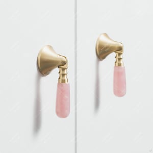 Peut inclure: Deux poignées de meuble en laiton et poignées en pierre rose. Les poignées ont un design incurvé avec une base décorative et une poignée lisse et allongée en pierre précieuse. Le matériel est monté sur une surface blanche.