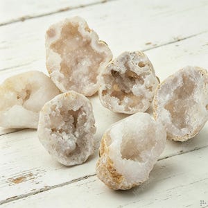 May include: A collection of six small, irregular geode stones featuring hollow centers lined with sparkling, translucent white quartz crystals. The outer surfaces of these natural mineral specimens are textured, matte, and light beige in color.