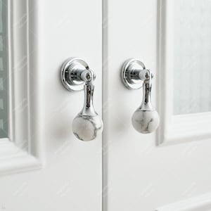 Op de afbeelding: Twee ronde deurknopjes van wit marmer en chroom. De knoppen zijn gemonteerd op een witte deur.