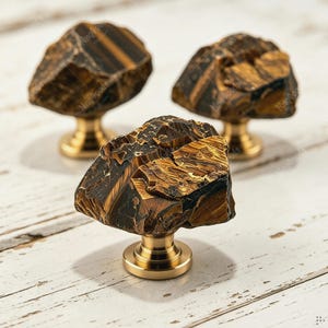 Puede incluir: Tres pomos para armarios con piedras de ojo de tigre en bruto y sin pulir, con bandas naturales en tonos marrón y dorado. Cada piedra está montada sobre una base metálica circular de color latón cepillado con un vástago cilíndrico corto.