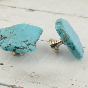 Puede incluir: Dos pomos para armarios con tapas de piedra turquesa plana e irregular con vetas naturales de color marrón oscuro. Cada piedra está montada sobre una base metálica pulida y escalonada diseñada para fijarse a muebles o puertas.