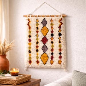 Puede incluir: Colgante de pared texturizado color crema con un patrón geométrico de diamantes en amarillo, rojo, negro y morado. La decoración de pared está suspendida de una clavija de madera con una cuerda y borlas.