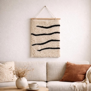 Puede incluir: Un tapiz de pared rectangular con una superficie texturizada de color crema y tres líneas onduladas negras. La obra de arte está suspendida de una barra de madera con una cuerda color crema.