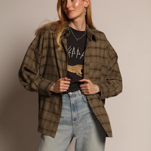Pode incluir: Uma mulher vestindo uma camisa de flanela xadrez verde escuro e marrom, uma camiseta preta com gráficos e jeans azul claro. A camiseta preta tem texto branco e um desenho de estampa de leopardo. A roupa é casual e estilosa.