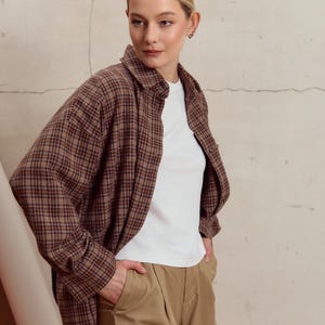 Pode incluir: Uma mulher vestindo uma camisa marrom e preta xadrez sobre uma camiseta branca e calças cáqui.