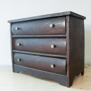 May include: A small, dark brown wooden chest of drawers featuring three horizontal drawers. Each drawer is equipped with two small, circular, metallic-colored knobs. The wood grain is visible throughout the structure, which has a rectangular top.