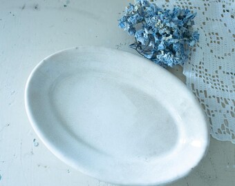 May include: An oval-shaped white ceramic dish with a smooth, glossy finish. Beside the dish lies a cluster of dried blue hydrangea flowers and a piece of white crocheted lace fabric with a detailed, intricate openwork pattern.
