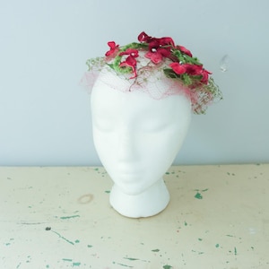 May include: A vintage floral hat with a white head form. The hat features a delicate net base, adorned with vibrant red flowers and green foliage. The head form is white and smooth, set against a light blue background.