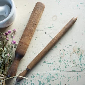 May include: Two wooden rolling pins, one long and thin, the other shorter and thicker, on a white wooden surface with green paint splatters. A white ceramic mortar and pestle is in the background.