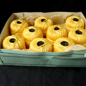 May include: A box filled with yellow spools of thread. The spools are arranged in a circular pattern and have a black label with the text "DMC" on them.