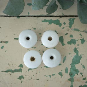 May include: Four white ceramic cabinet knobs with a circular design and a central hole. The knobs are arranged on a distressed, cream-colored surface with green paint accents. Eucalyptus leaves are in the background.