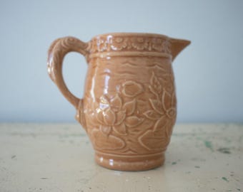 May include: A tan ceramic pitcher with a detailed floral design. The pitcher has a curved handle and a spout for pouring. The floral pattern is embossed on the body of the pitcher, adding texture and visual interest. The pitcher is likely used for serving beverages.