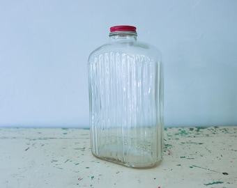 May include: A clear glass bottle with a rectangular shape and vertical ribbed texture along the sides. The bottle features a narrow neck topped with a solid red metal screw-on cap. The glass is transparent and smooth, showcasing a simple design.