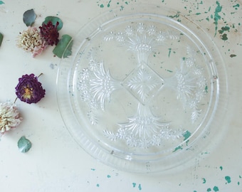 May include: A clear, round glass cake stand with a decorative floral pattern. The stand is surrounded by dried flowers in shades of purple, cream, and burgundy, along with green leaves. The stand is on a white surface.